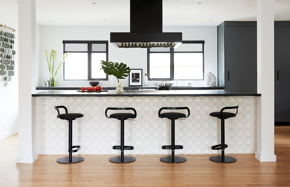 This is an example of a large contemporary single-wall open plan kitchen in San Francisco with an undermount sink, flat-panel cabinets, black cabinets, soapstone benchtops, white splashback, porcelain splashback, stainless steel appliances, light hardwood floors, with island, brown floor and black benchtop.