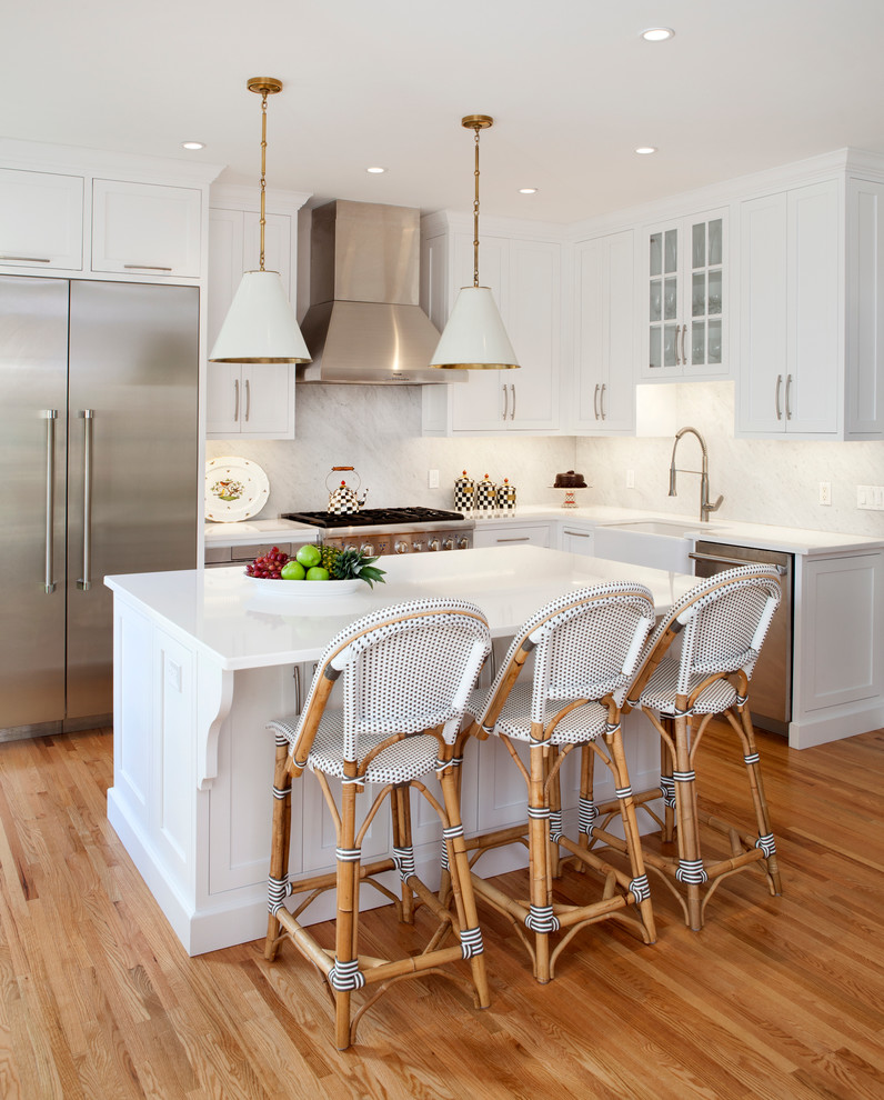 Photo of a transitional l-shaped kitchen in New York with a farmhouse sink, shaker cabinets, white cabinets, white splashback, medium hardwood floors, with island, marble benchtops, marble splashback, stainless steel appliances and white benchtop.