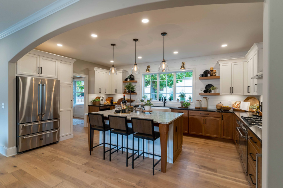 Inspiration for a large country u-shaped open plan kitchen in Other with a farmhouse sink, recessed-panel cabinets, medium wood cabinets, granite benchtops, white splashback, porcelain splashback, stainless steel appliances, light hardwood floors, with island, brown floor and black benchtop.