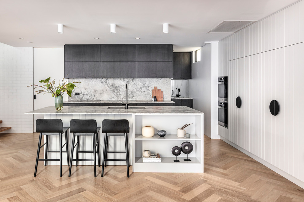 Photo of a beach style l-shaped kitchen in Sydney with black cabinets, marble benchtops, with island, an undermount sink, white splashback, black appliances, light hardwood floors, beige floor and white benchtop.
