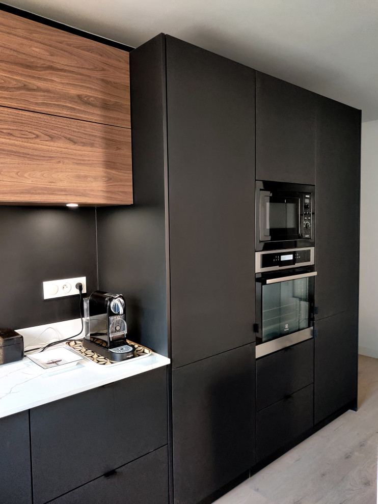 Photo of a mid-sized contemporary l-shaped open plan kitchen in Lille with a single-bowl sink, beaded inset cabinets, black cabinets, quartzite benchtops, white splashback, engineered quartz splashback, black appliances, light hardwood floors, with island and white benchtop.
