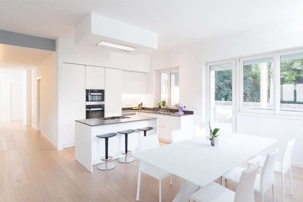 This is an example of a large contemporary l-shaped open plan kitchen in Other with a single-bowl sink, flat-panel cabinets, white cabinets, white splashback, black appliances, light hardwood floors and with island.