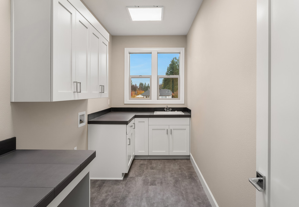 Inspiration for a mid-sized transitional l-shaped dedicated laundry room in Portland with a drop-in sink, recessed-panel cabinets, white cabinets, tile benchtops, grey walls, vinyl floors, a side-by-side washer and dryer, grey floor and black benchtop.