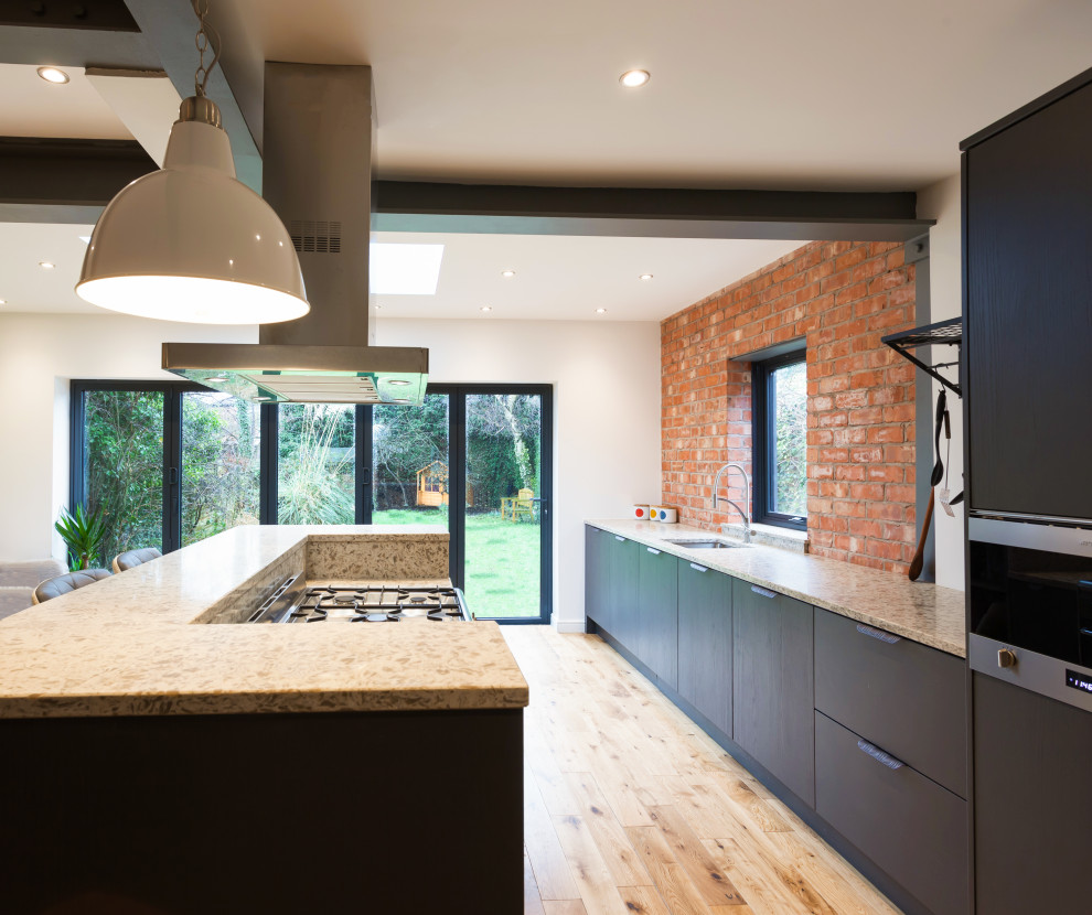Design ideas for a large industrial single-wall open plan kitchen in Cheshire with an undermount sink, flat-panel cabinets, grey cabinets, quartzite benchtops, multi-coloured splashback, brick splashback, stainless steel appliances, light hardwood floors, with island and grey benchtop.