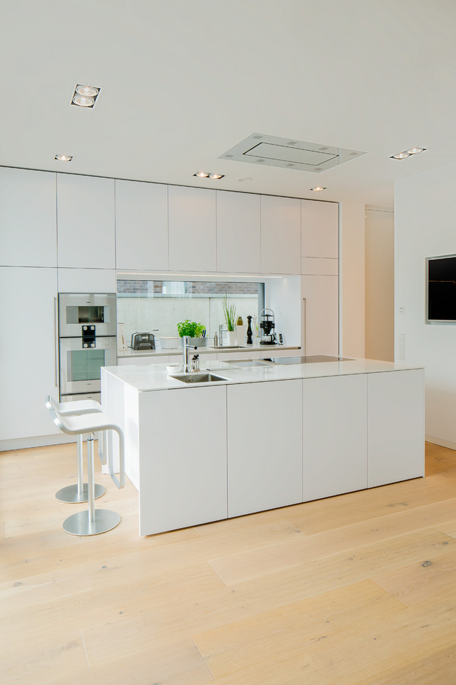 Inspiration for a large modern galley open plan kitchen in Dusseldorf with an undermount sink, flat-panel cabinets, white cabinets, solid surface benchtops, white splashback, glass sheet splashback, white appliances, medium hardwood floors, with island, brown floor and white benchtop.