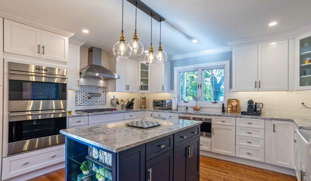 This is an example of a mid-sized transitional u-shaped eat-in kitchen in Philadelphia with a farmhouse sink, shaker cabinets, blue cabinets, quartz benchtops, white splashback, porcelain splashback, stainless steel appliances, medium hardwood floors, with island, brown floor and grey benchtop.