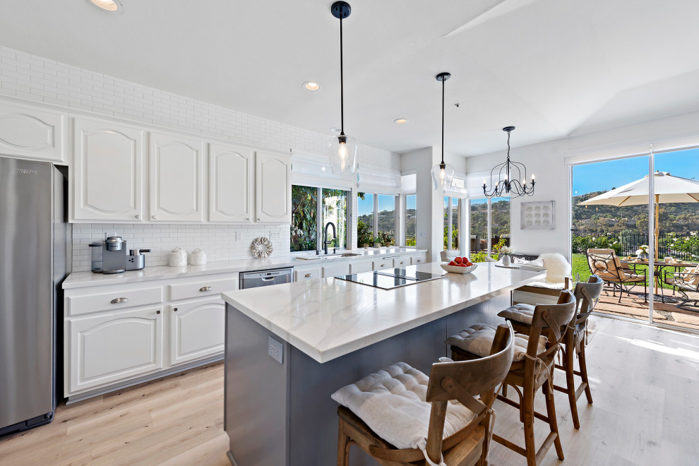 Photo of a mid-sized beach style single-wall eat-in kitchen in Orange County with an undermount sink, raised-panel cabinets, white cabinets, quartz benchtops, white splashback, ceramic splashback, stainless steel appliances, vinyl floors, with island, beige floor and white benchtop.