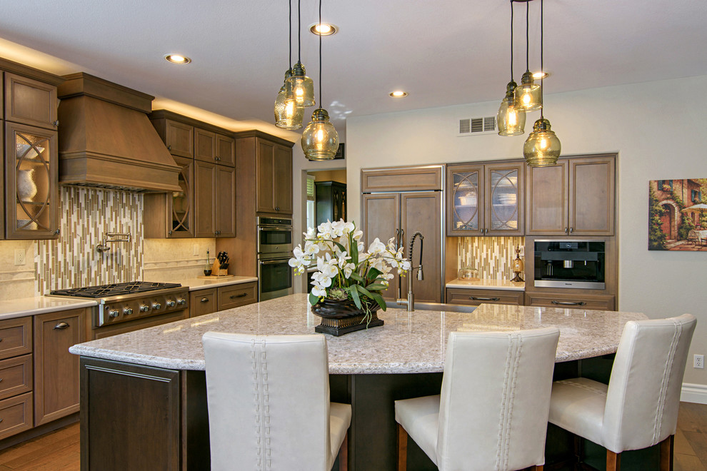 Example of a large transitional l-shaped medium tone wood floor and brown floor open concept kitchen design in San Diego with a farmhouse sink, recessed-panel cabinets, medium tone wood cabinets, beige backsplash, paneled appliances, an island, quartz countertops and travertine backsplash
