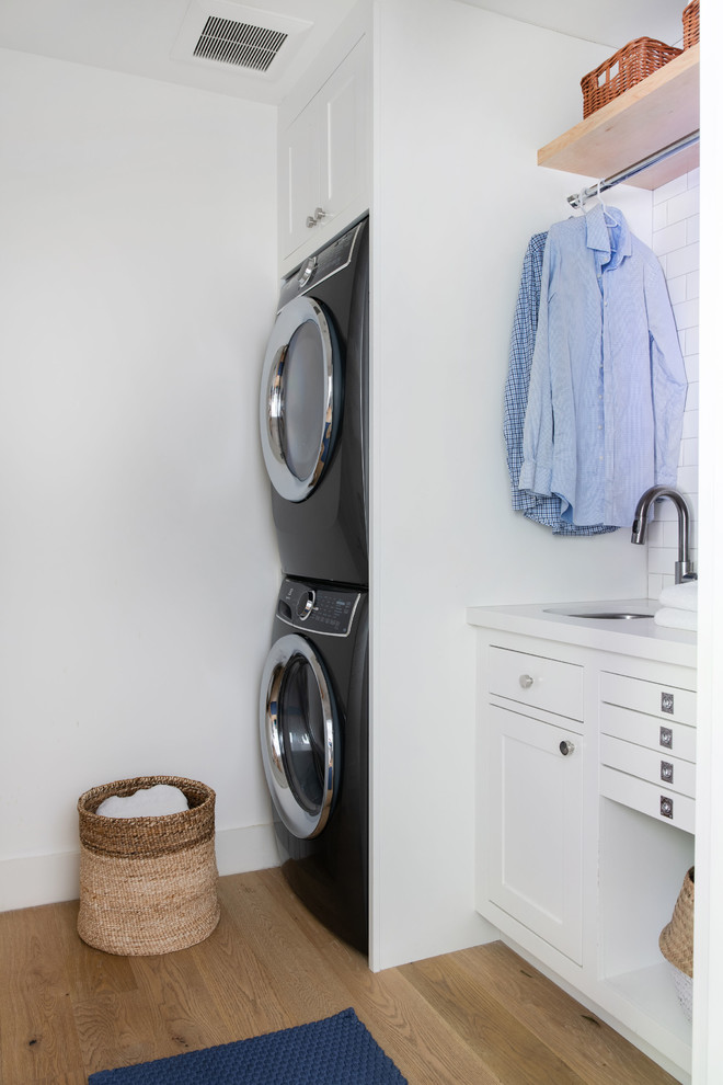 Small transitional dedicated laundry room in Los Angeles with an undermount sink, shaker cabinets, white cabinets, quartz benchtops, white walls, medium hardwood floors, a stacked washer and dryer, brown floor and white benchtop.