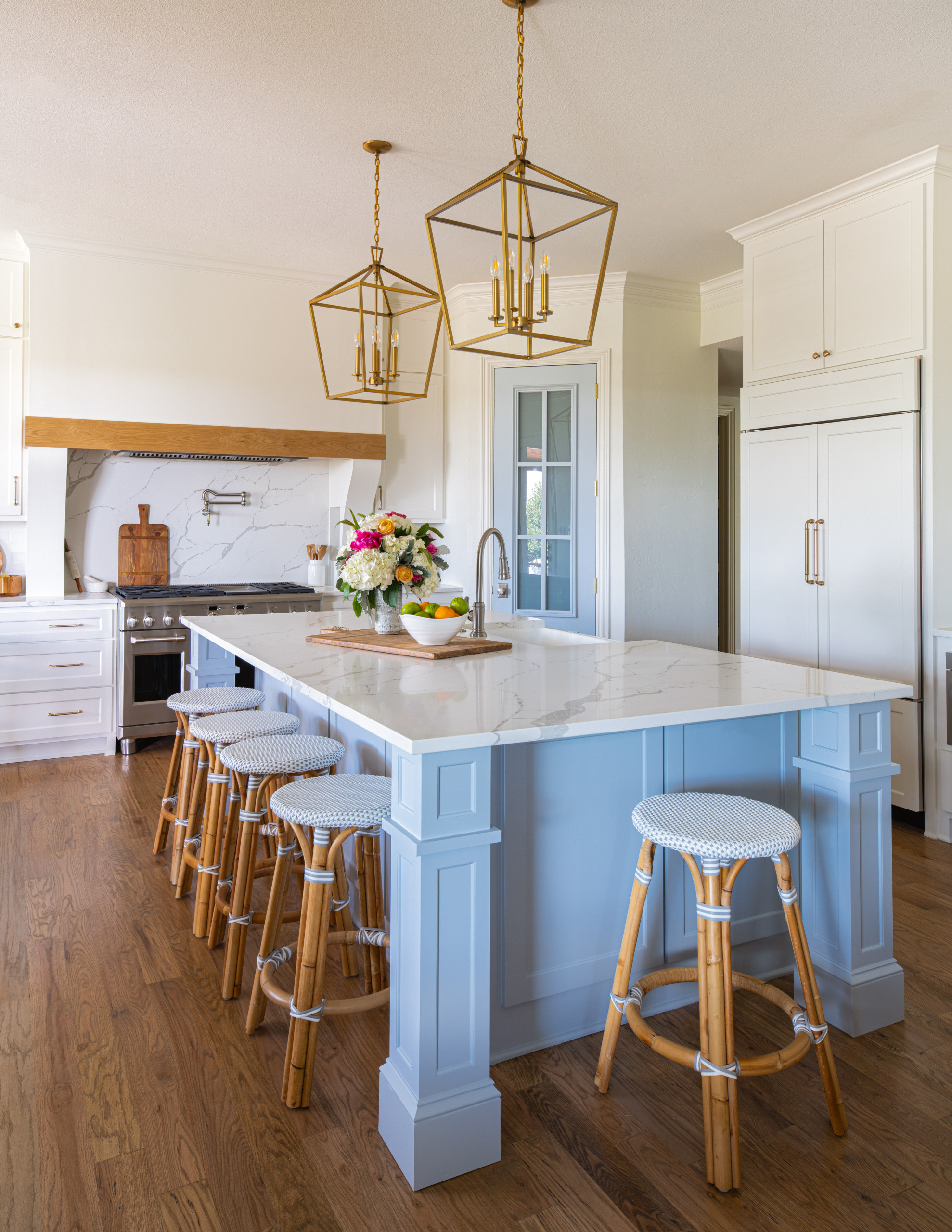 White transitional kitchen with large vent hood with white oak accent, blue island, blue pantry door, brass lighting, brass hardware, cast iron apron front sink.