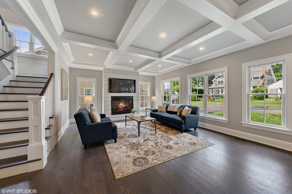 This is an example of a mid-sized transitional open concept family room in Chicago with grey walls, dark hardwood floors, a standard fireplace, a wood fireplace surround, a wall-mounted tv and brown floor.