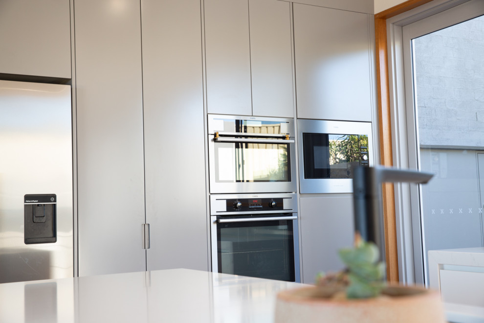 Beach style galley kitchen pantry in Central Coast with an undermount sink, white cabinets, grey splashback, glass sheet splashback, black appliances, light hardwood floors, with island, brown floor and white benchtop.