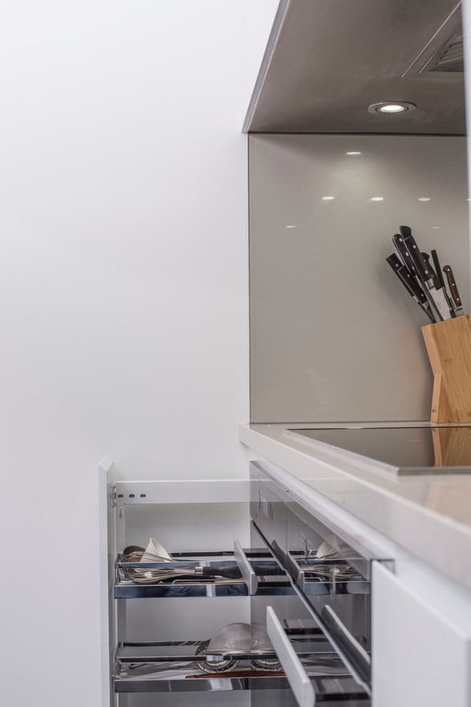 This is an example of a mid-sized scandinavian l-shaped separate kitchen in Brisbane with an undermount sink, flat-panel cabinets, white cabinets, quartz benchtops, metallic splashback, glass sheet splashback, black appliances, porcelain floors, grey floor and grey benchtop.