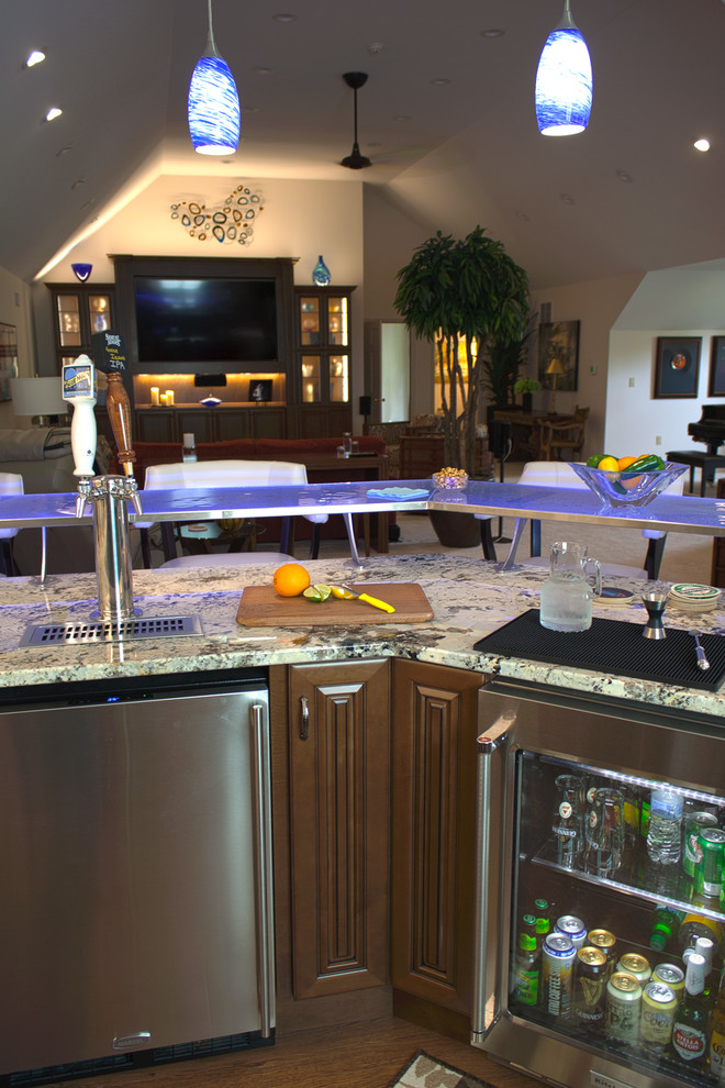 This is an example of a large transitional l-shaped seated home bar in Huntington with an undermount sink, glass-front cabinets, dark wood cabinets, glass benchtops, medium hardwood floors and brown floor.