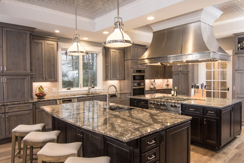 Photo of a large traditional open plan kitchen in Denver with an undermount sink, raised-panel cabinets, stainless steel benchtops, white splashback, stone tile splashback, stainless steel appliances, light hardwood floors, multiple islands, beige benchtop and coffered.