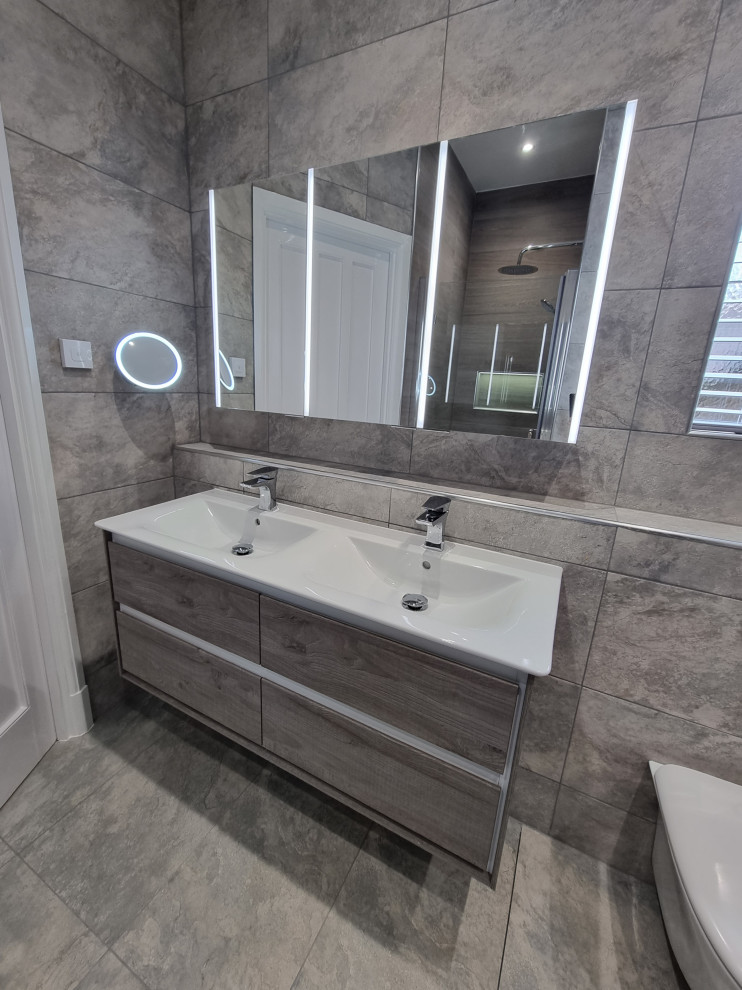 Large contemporary grey and brown ensuite bathroom in West Midlands with flat-panel cabinets, grey cabinets, a freestanding bath, a built-in shower, a wall mounted toilet, grey tiles, porcelain tiles, grey walls, porcelain flooring, a wall-mounted sink, grey floors, a hinged door, a wall niche, double sinks and a floating vanity unit.