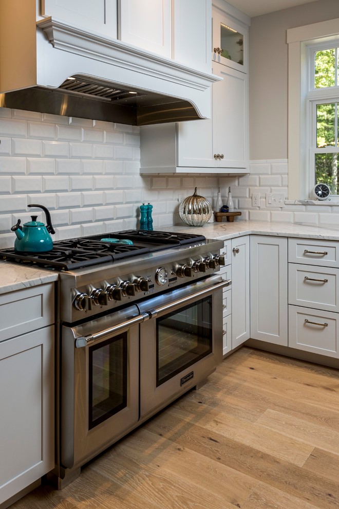 This is an example of a large arts and crafts u-shaped open plan kitchen in Portland Maine with a farmhouse sink, recessed-panel cabinets, white cabinets, quartz benchtops, white splashback, subway tile splashback, stainless steel appliances, light hardwood floors and with island.