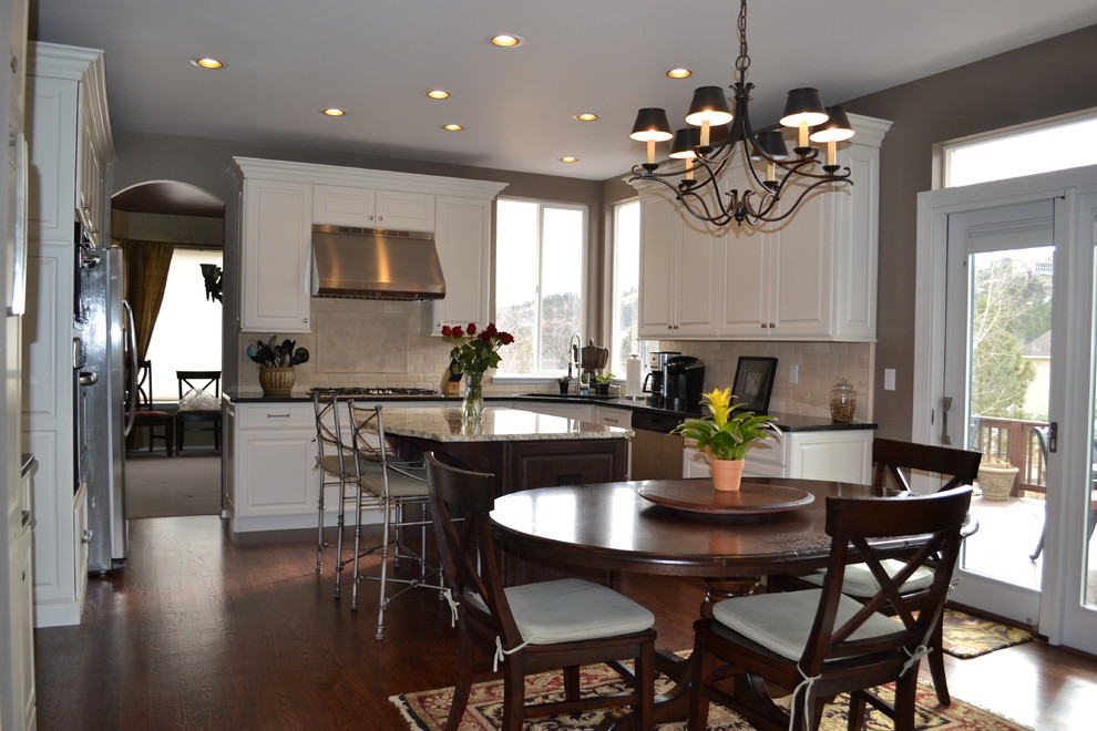 Example of a mid-sized mountain style u-shaped medium tone wood floor eat-in kitchen design in Denver with an undermount sink, raised-panel cabinets, white cabinets, granite countertops, beige backsplash, stone tile backsplash, stainless steel appliances and an island