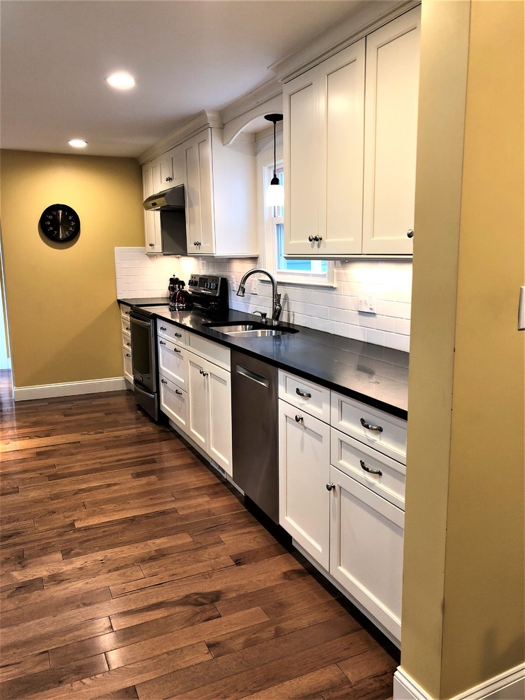 Small transitional galley medium tone wood floor and brown floor enclosed kitchen photo in Boston with an undermount sink, shaker cabinets, white cabinets, granite countertops, white backsplash, subway tile backsplash, stainless steel appliances and black countertops