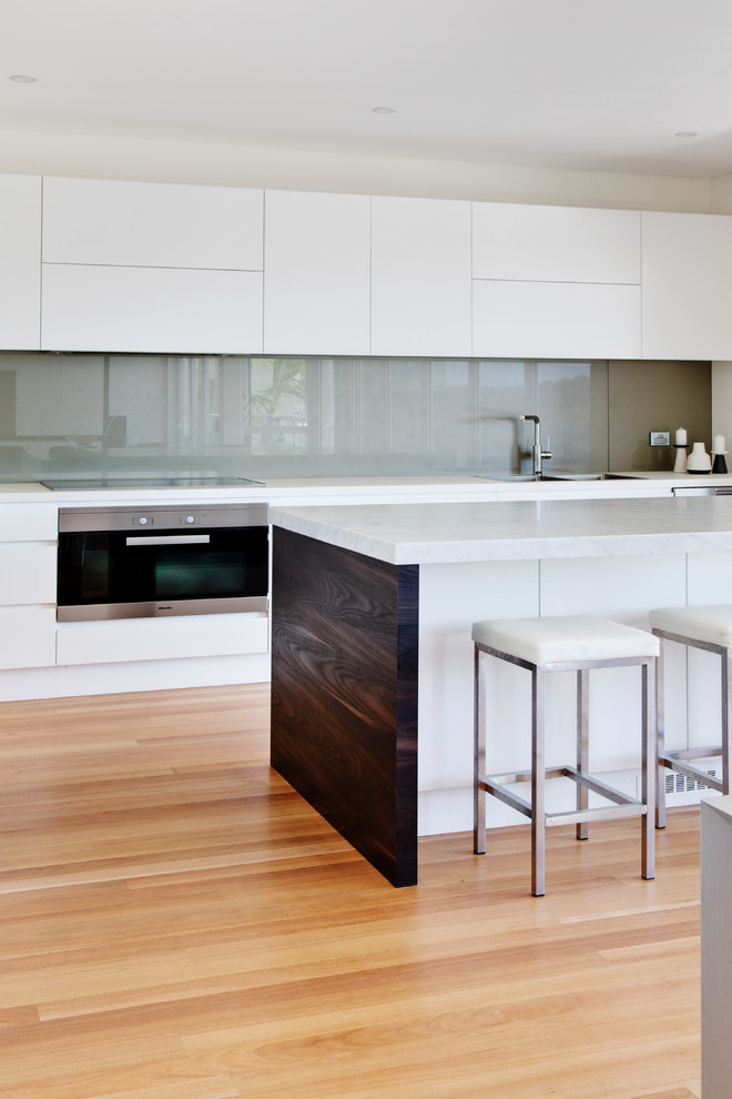 Design ideas for a mid-sized modern single-wall open plan kitchen in Sydney with a double-bowl sink, flat-panel cabinets, white cabinets, marble benchtops, grey splashback, glass sheet splashback, stainless steel appliances, light hardwood floors, with island, brown floor and grey benchtop.
