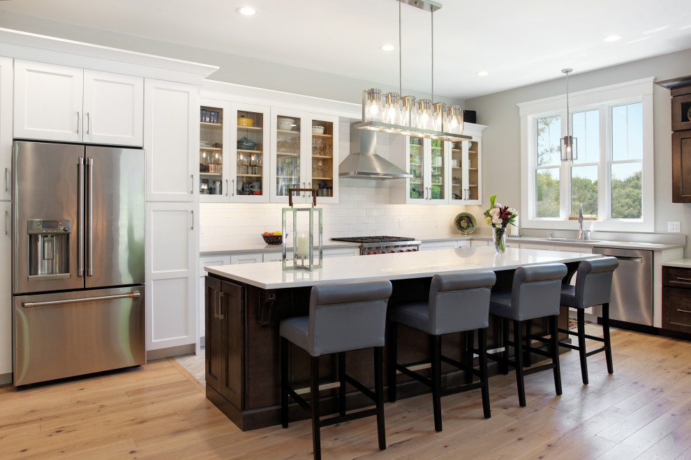 Example of a mid-sized transitional l-shaped light wood floor open concept kitchen design in Other with a drop-in sink, beaded inset cabinets, white cabinets, quartz countertops, white backsplash, ceramic backsplash, stainless steel appliances, an island and white countertops