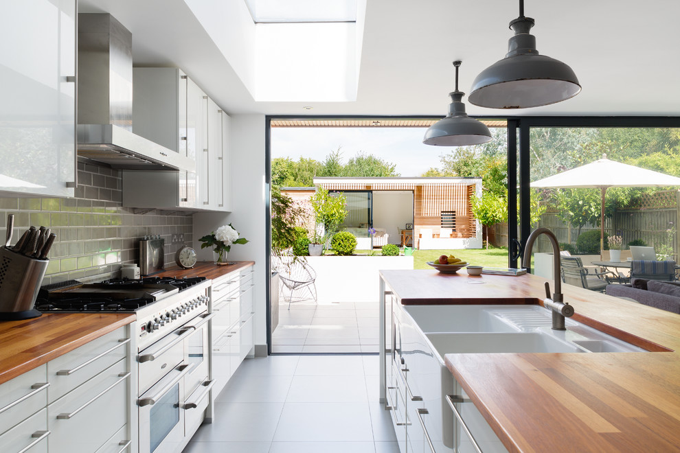 Design ideas for a mid-sized contemporary galley open plan kitchen in London with a farmhouse sink, flat-panel cabinets, white cabinets, wood benchtops, grey splashback, subway tile splashback and with island.