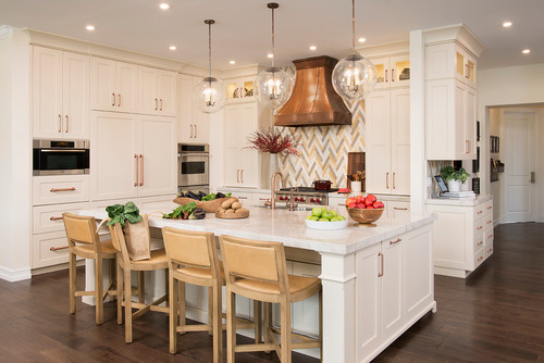 Large white kitchen with pops of copper, browns, tan, with pendant lighting, four bar stools, and a copper hood.