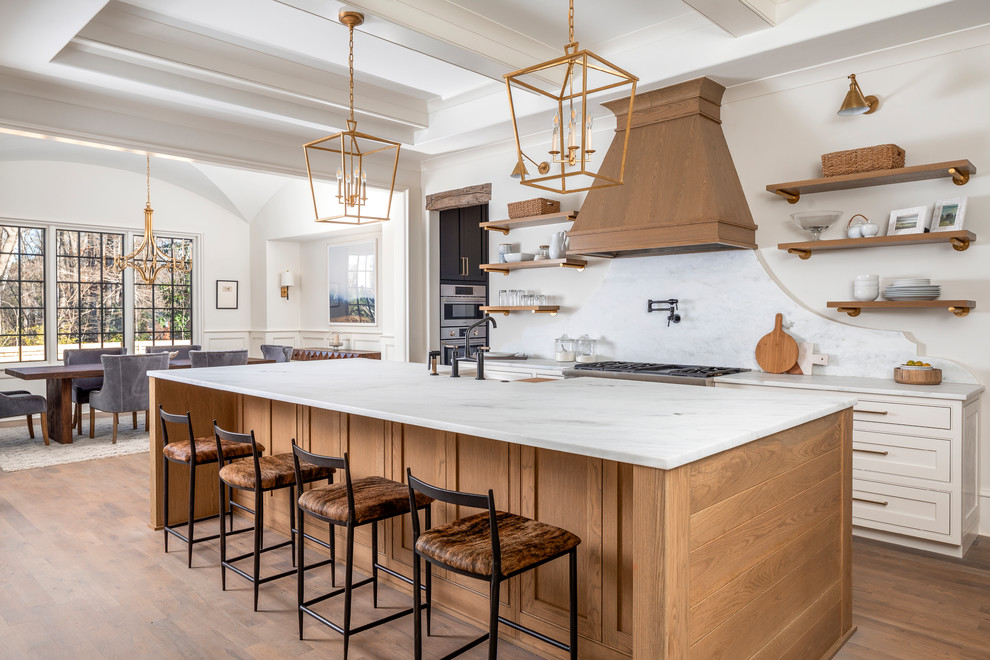 This is an example of a beach style eat-in kitchen in Charlotte with shaker cabinets, white cabinets, white splashback, stone slab splashback, stainless steel appliances, light hardwood floors, with island and white benchtop.