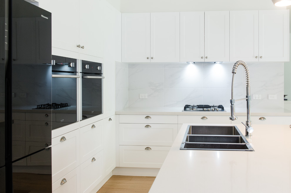 Large scandinavian l-shaped kitchen pantry in Melbourne with a double-bowl sink, shaker cabinets, white cabinets, solid surface benchtops, white splashback, ceramic splashback, black appliances, light hardwood floors and with island.