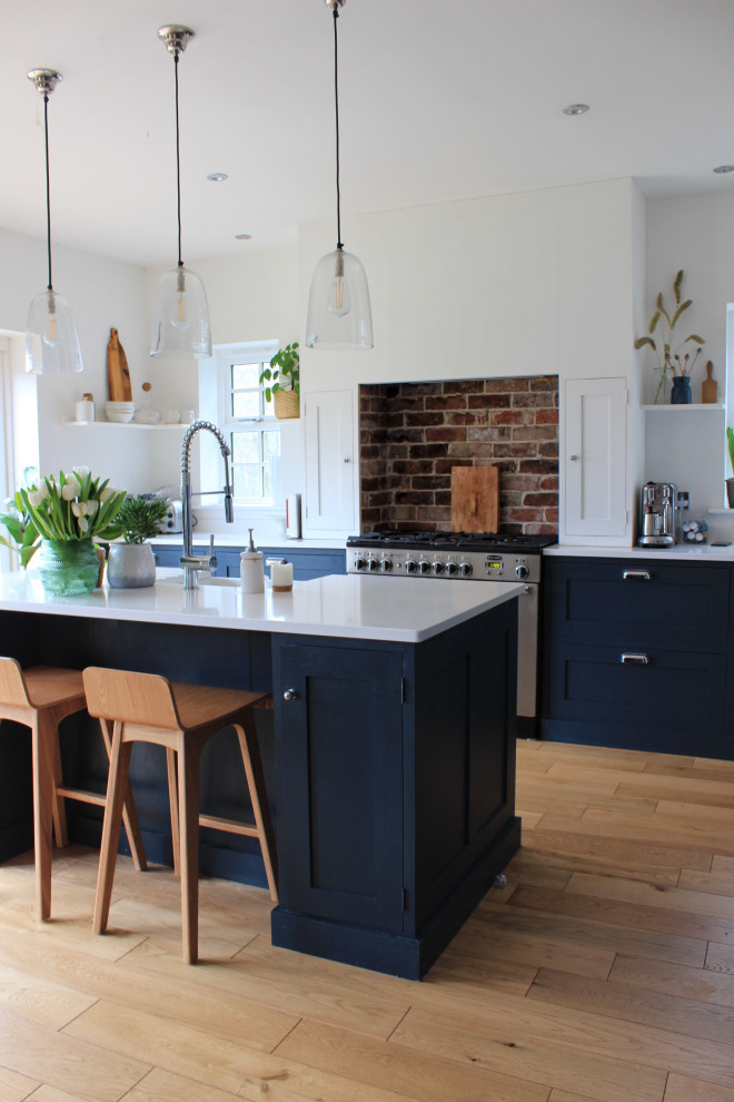 This is an example of a large contemporary single-wall eat-in kitchen in West Midlands with a farmhouse sink, shaker cabinets, blue cabinets, quartzite benchtops, brick splashback, panelled appliances, medium hardwood floors, with island and white benchtop.