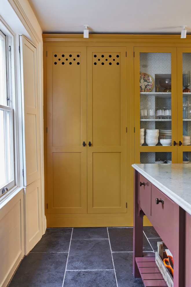 Inspiration for a large traditional kitchen/diner in London with a belfast sink, shaker cabinets, yellow cabinets, marble worktops, white splashback, ceramic splashback, white appliances, limestone flooring, an island, black floors, brown worktops and a chimney breast.