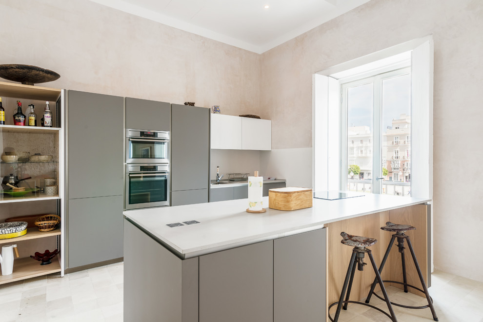 This is an example of a mediterranean kitchen in Other with flat-panel cabinets, grey cabinets, white splashback, stainless steel appliances, with island, white floor and white benchtop.