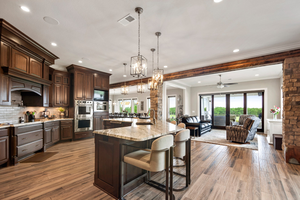 Expansive country single-wall open plan kitchen in Kansas City with an undermount sink, raised-panel cabinets, dark wood cabinets, marble benchtops, beige splashback, stone tile splashback, stainless steel appliances, ceramic floors, with island, brown floor and brown benchtop.