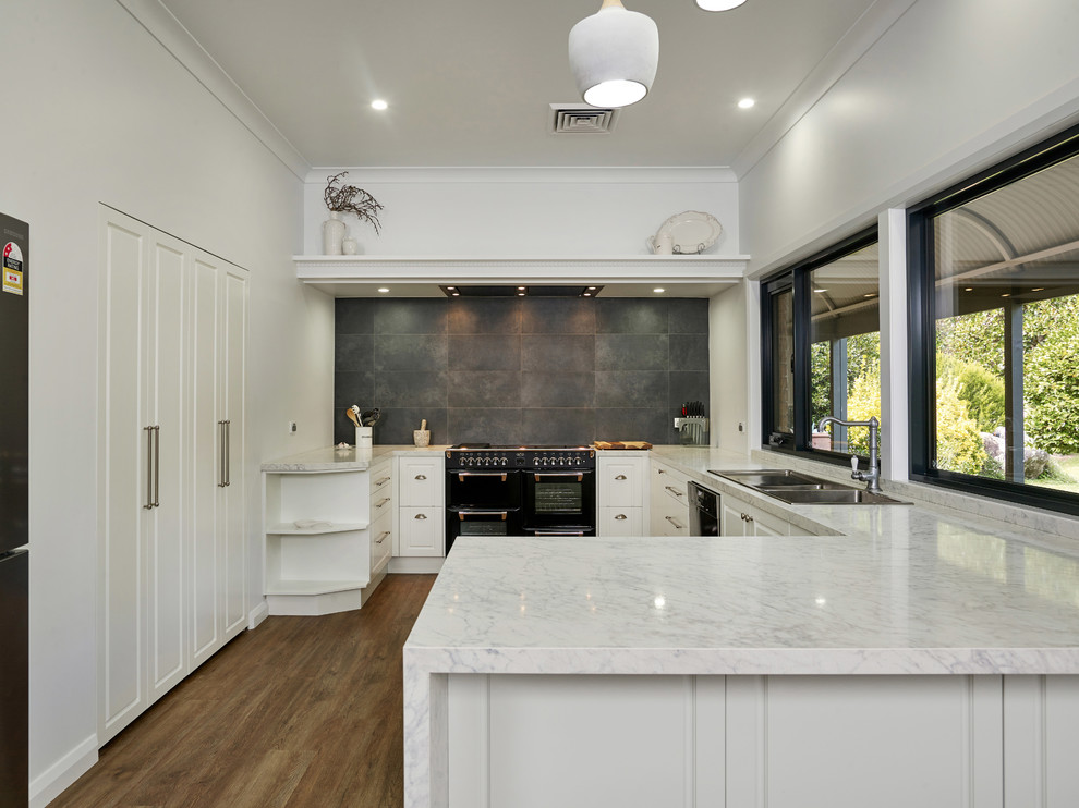 Inspiration for a mid-sized u-shaped kitchen pantry with a drop-in sink, recessed-panel cabinets, white cabinets, quartz benchtops, grey splashback, ceramic splashback, black appliances, medium hardwood floors, with island, brown floor and white benchtop.