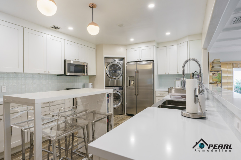 This is an example of a large modern u-shaped kitchen/diner in Los Angeles with a belfast sink, shaker cabinets, white cabinets, composite countertops, white splashback, stone tiled splashback, stainless steel appliances, light hardwood flooring, an island, brown floors and white worktops.