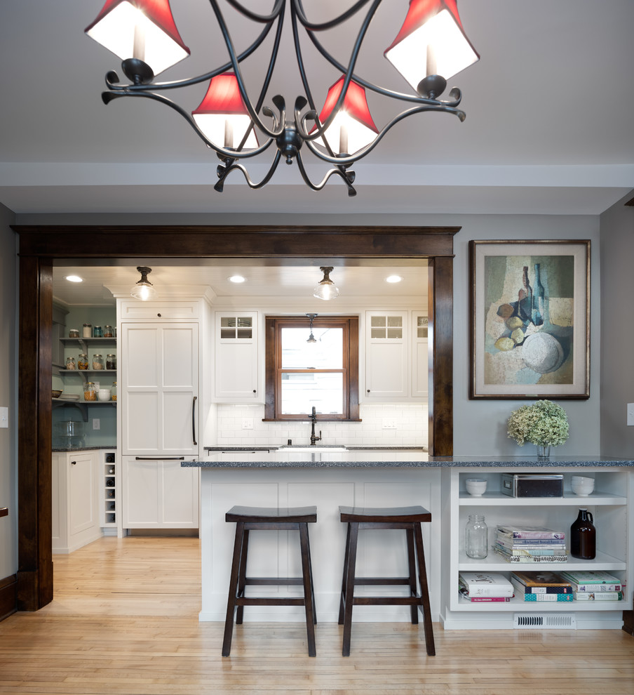 Photo of a mid-sized traditional u-shaped eat-in kitchen in Minneapolis with a farmhouse sink, shaker cabinets, white cabinets, recycled glass benchtops, white splashback, subway tile splashback, panelled appliances, light hardwood floors, a peninsula, brown floor and grey benchtop.