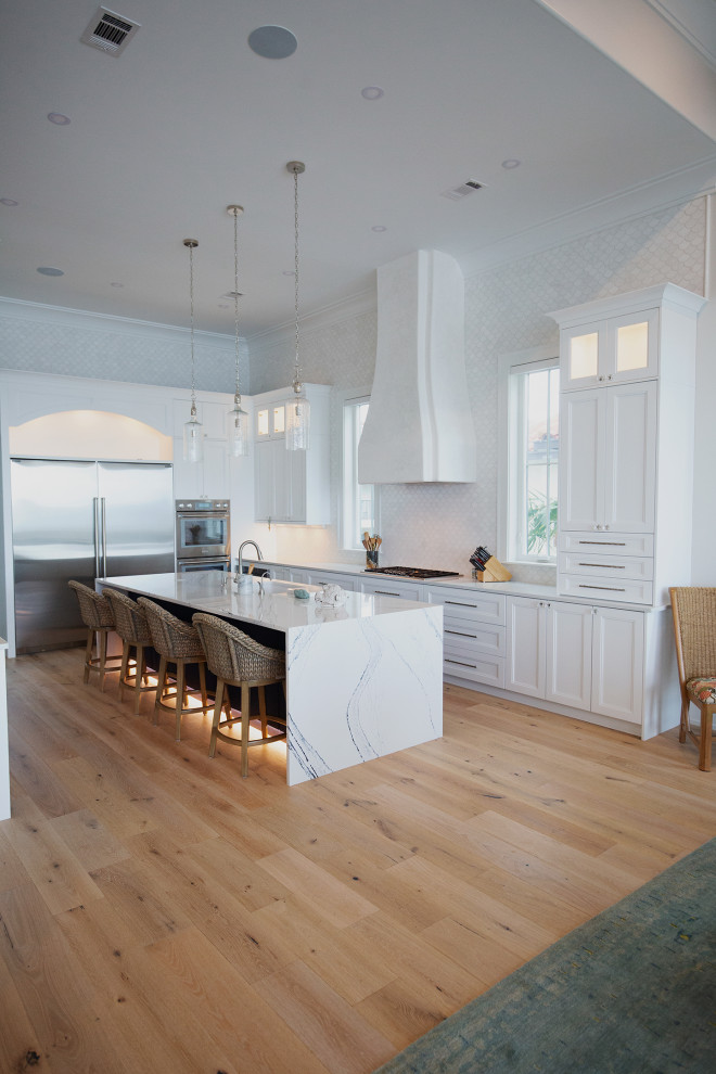 Photo of an expansive beach style l-shaped eat-in kitchen in Miami with a drop-in sink, shaker cabinets, white cabinets, quartzite benchtops, white splashback, ceramic splashback, stainless steel appliances, medium hardwood floors, with island, beige floor and white benchtop.