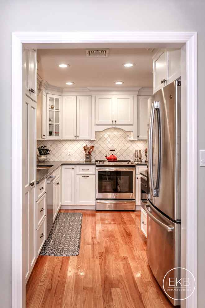 Photo of a small traditional galley separate kitchen in New York with an undermount sink, recessed-panel cabinets, white cabinets, soapstone benchtops, white splashback, ceramic splashback, stainless steel appliances, light hardwood floors, no island, brown floor and grey benchtop.