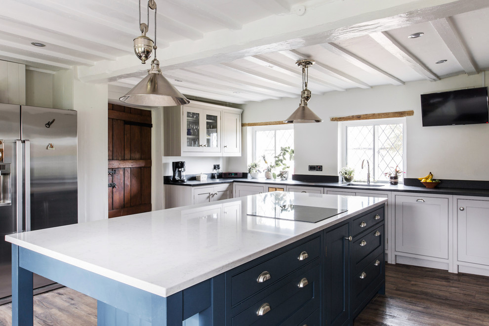 This is an example of a large country u-shaped open plan kitchen in Kent with an integrated sink, shaker cabinets, white cabinets, granite benchtops, black appliances, medium hardwood floors, with island and black benchtop.