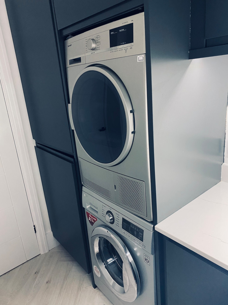 Photo of a small beach style l-shaped utility room in Other with a submerged sink, flat-panel cabinets, blue cabinets, quartz worktops, white splashback, engineered quartz splashback, white walls, laminate floors, a stacked washer and dryer, black floors and white worktops.