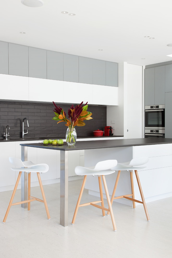 Photo of a mid-sized modern open plan kitchen in Melbourne with flat-panel cabinets, white cabinets, stainless steel appliances, with island, white floor, a double-bowl sink, black splashback, ceramic splashback, cork floors and black benchtop.