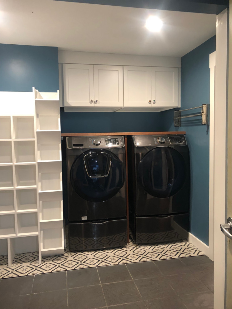 This is an example of an eclectic single-wall utility room in Boston with shaker cabinets, white cabinets, wood benchtops, blue walls, ceramic floors, a side-by-side washer and dryer and grey floor.