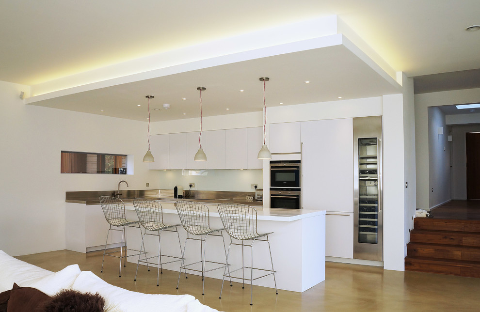 Photo of a large contemporary u-shaped open plan kitchen in Surrey with a drop-in sink, flat-panel cabinets, white cabinets, solid surface benchtops, metallic splashback, metal splashback, stainless steel appliances, laminate floors and a peninsula.