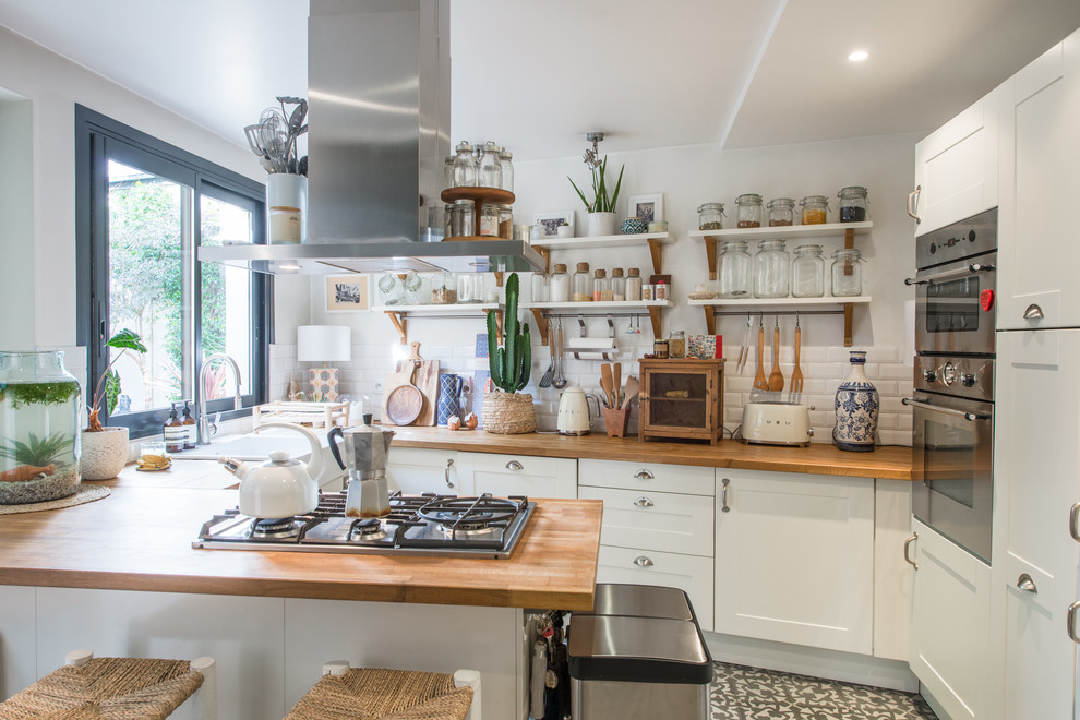 Inspiration for an eclectic u-shaped kitchen in Paris with a farmhouse sink, shaker cabinets, white cabinets, wood benchtops, white splashback, subway tile splashback, stainless steel appliances, a peninsula and grey floor.