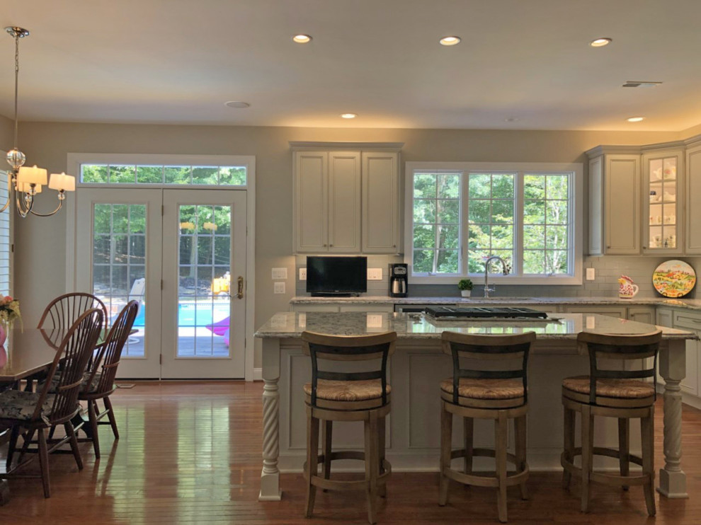 Photo of a large traditional l-shaped eat-in kitchen in Baltimore with a double-bowl sink, recessed-panel cabinets, white cabinets, quartz benchtops, grey splashback, subway tile splashback, stainless steel appliances, medium hardwood floors, with island, brown floor and grey benchtop.