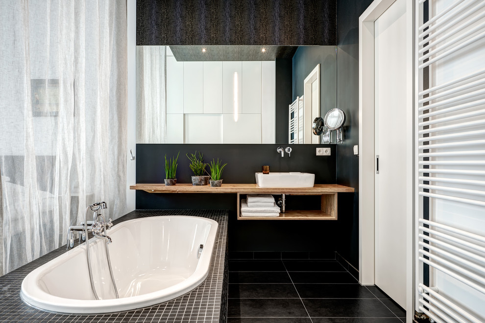 Photo of a small contemporary master bathroom in Berlin with open cabinets, brown cabinets, a drop-in tub, black walls, a vessel sink, wood benchtops, black floor and brown benchtops.