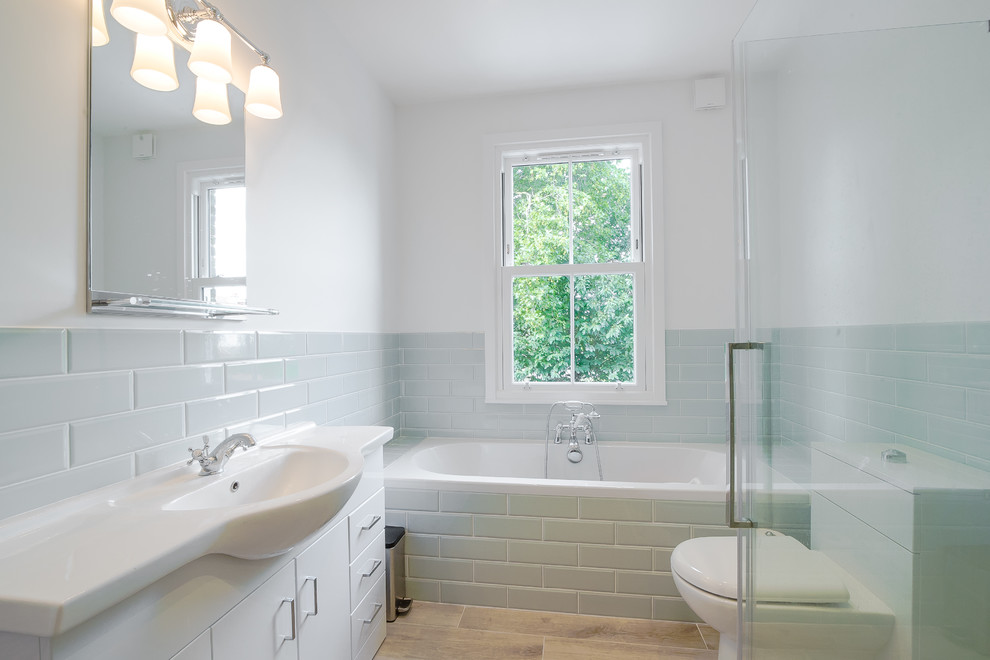 Small transitional kids bathroom in London with flat-panel cabinets, white cabinets, a drop-in tub, gray tile, ceramic tile, white walls, porcelain floors, brown floor, a hinged shower door and a console sink.