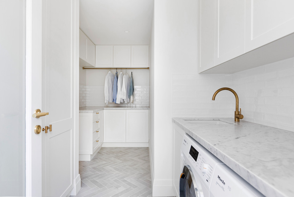 This is an example of a mid-sized beach style l-shaped dedicated laundry room in Sydney with a drop-in sink, shaker cabinets, white cabinets, marble benchtops, white splashback, subway tile splashback, white walls, ceramic floors, a side-by-side washer and dryer, grey floor and grey benchtop.