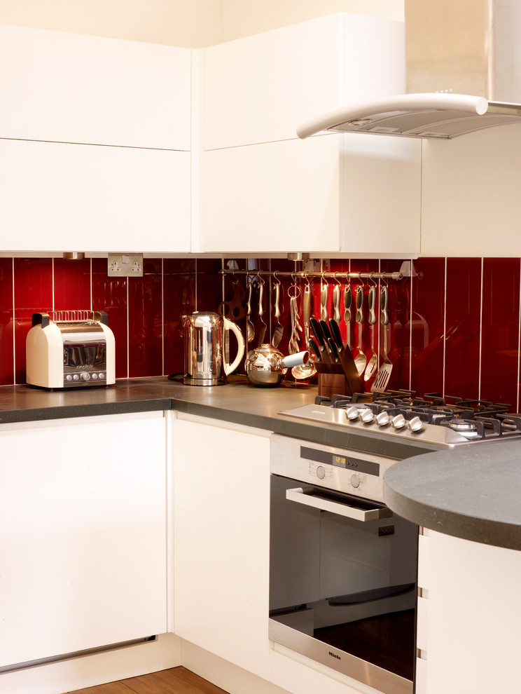 This is an example of a contemporary u-shaped open plan kitchen in London with flat-panel cabinets, white cabinets, red splashback, medium hardwood floors, a peninsula, wood benchtops and panelled appliances.