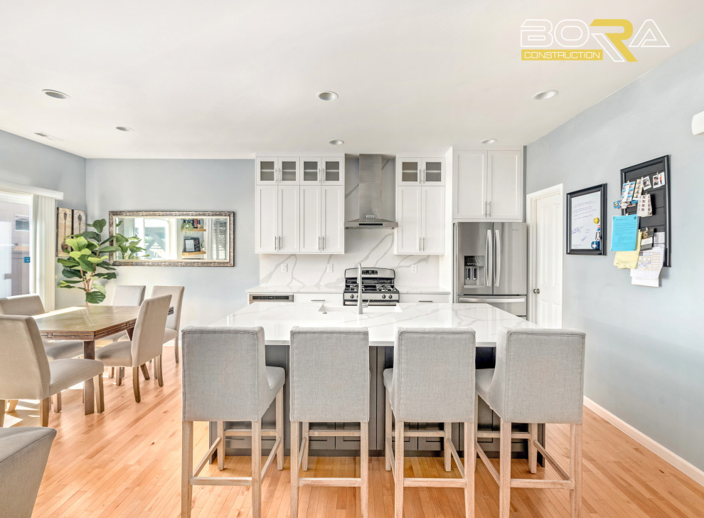 Mid-sized single-wall light wood floor and brown floor eat-in kitchen photo in Seattle with a farmhouse sink, shaker cabinets, white cabinets, quartz countertops, white backsplash, quartz backsplash, stainless steel appliances, an island and white countertops